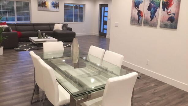 A modern living room with a glass-top dining table, white chairs, a black sofa, and a large window with blinds. There are three abstract paintings on the wall.
