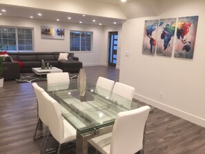 Una sala de estar moderna con una mesa de comedor de vidrio, sillas blancas, un sofá negro y un ventanal con persianas. Hay tres cuadros abstractos en la pared.