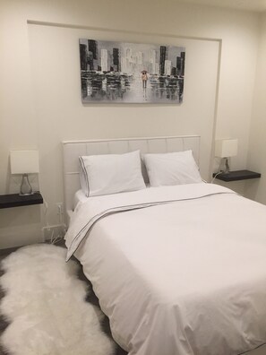 A neatly arranged bedroom with a large bed, white bedding, two bedside tables with lamps, a fluffy white rug, and a cityscape painting on the wall.