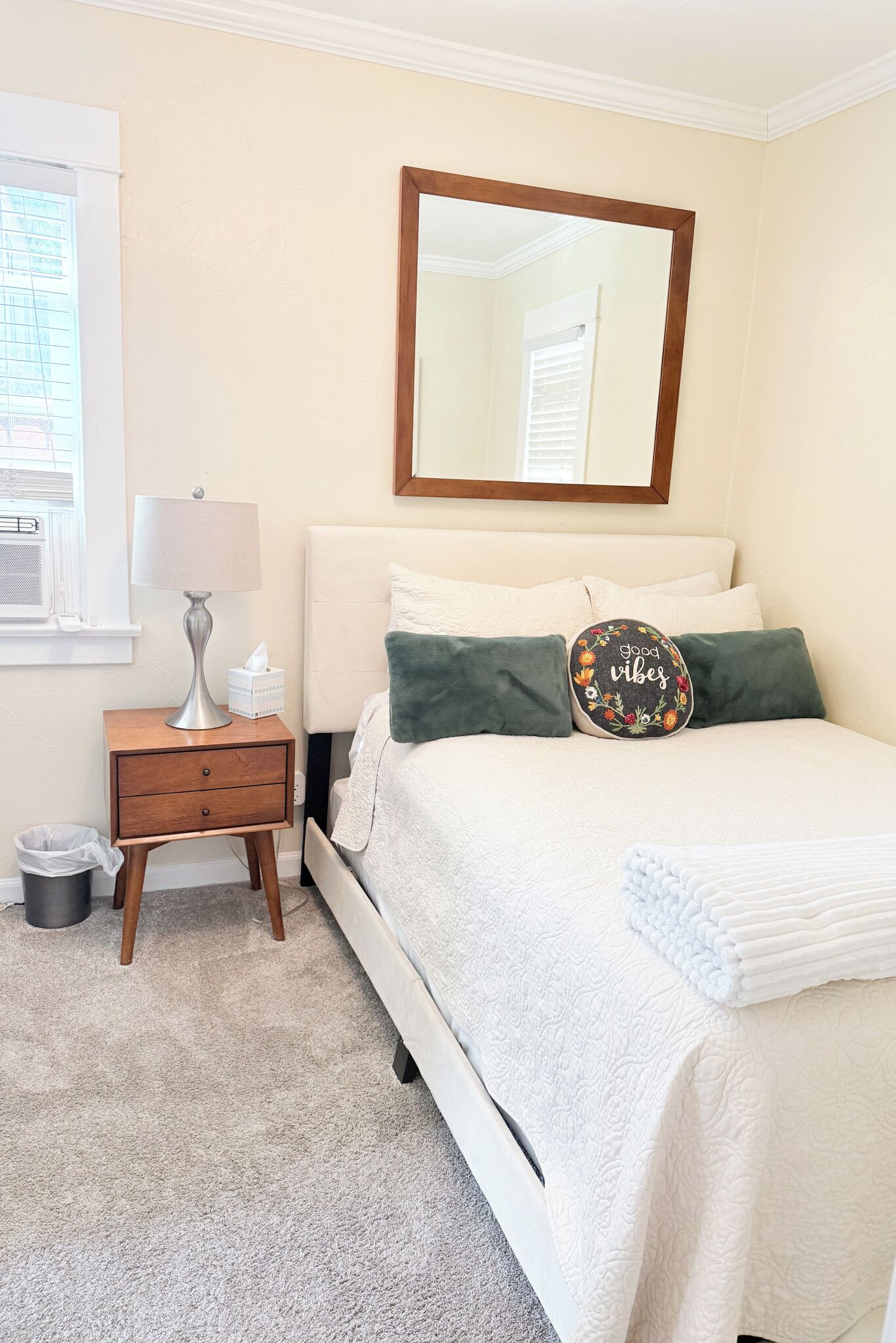 A bedroom with a bed, a nightstand, a lamp, a mirror, and a window.