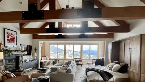 A spacious living room with a fireplace, large windows, and a high ceiling with exposed beams. - Beautiful Ski-In/Ski-Out Penthouse (Whitefish)