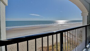 Une vue sur la plage depuis un balcon doté d’une balustrade, avec l’océan et un ciel dégagé en toile de fond.