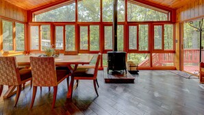 A spacious room with large windows, a central wood-burning stove, and a dining area with chairs and a table.