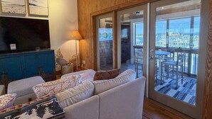 Een gezellige woonkamer met een bank, een televisie en een lamp. Door grote glazen deuren is er uitzicht op een jachthaven.