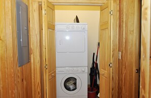 A laundry room with a stacked washer and dryer, wooden walls, and a red bucket. - Down Creek Condo 102: Luxurious, reverse floor plan, in the Village. (Ocracoke)