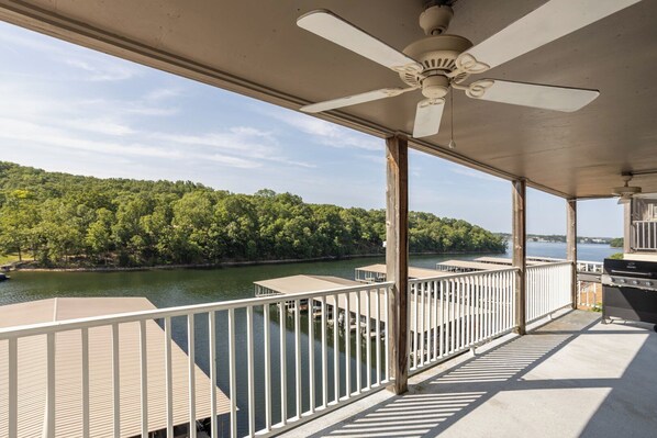 A covered deck with a ceiling fan, wooden railings, and a view of a lake and forest.