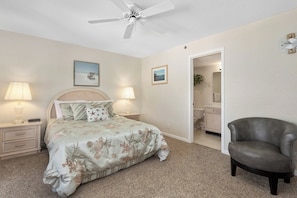 A bedroom with a bed, two bedside lamps, a ceiling fan, a chair, and a bathroom visible through an open door.