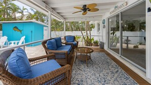 Un espace extérieur aménagé, abrité et doté de mobilier en osier et d’un ventilateur de plafond, offrant une vue sur une piscine et des palmiers.