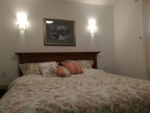 Un lit avec une literie à motifs floraux, deux lampes fixées au mur et un tableau encadré au-dessus.
