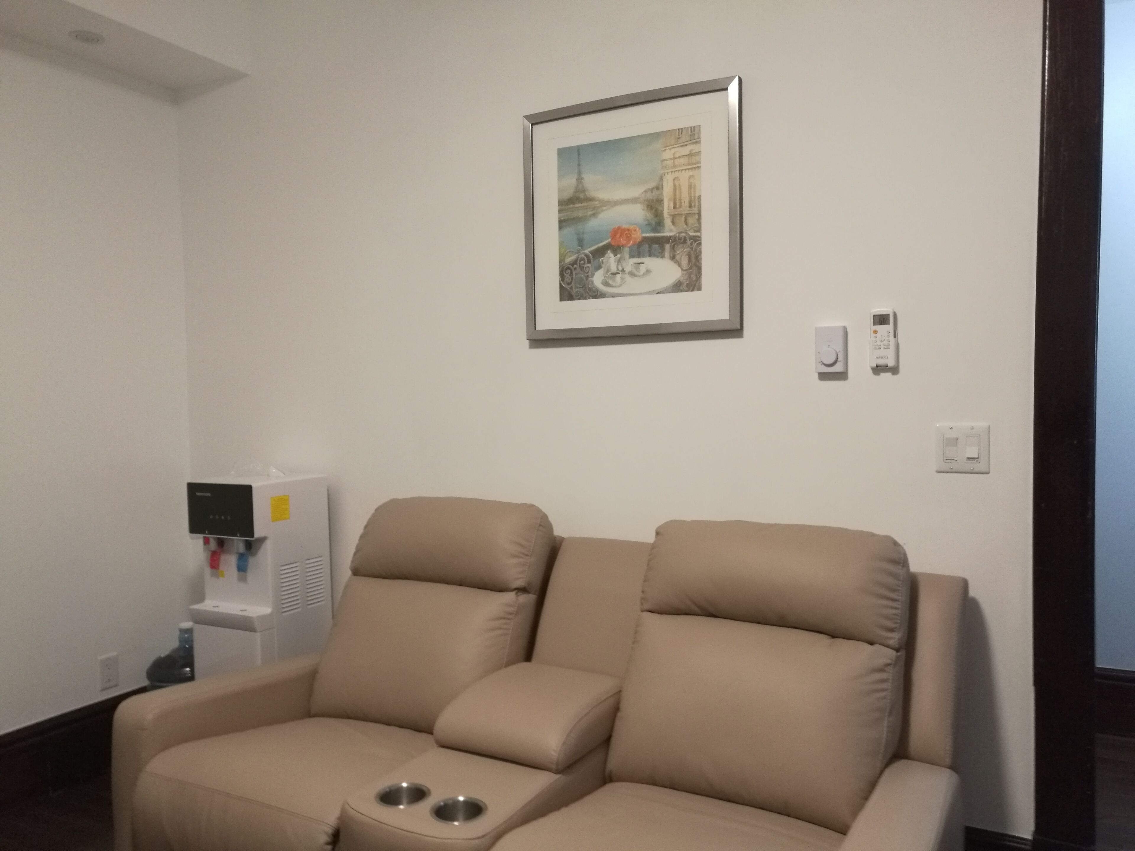 A room with a beige sofa, a water dispenser, a framed picture of a cityscape, and a thermostat on the wall.