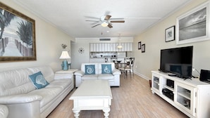 A spacious living room with a white sofa, a coffee table, a television, and a kitchen in the background.