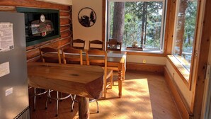Een rustiek houten interieur met een eettafel en stoelen, een koelkast, een plafondventilator en een groot raam met uitzicht op het bos.