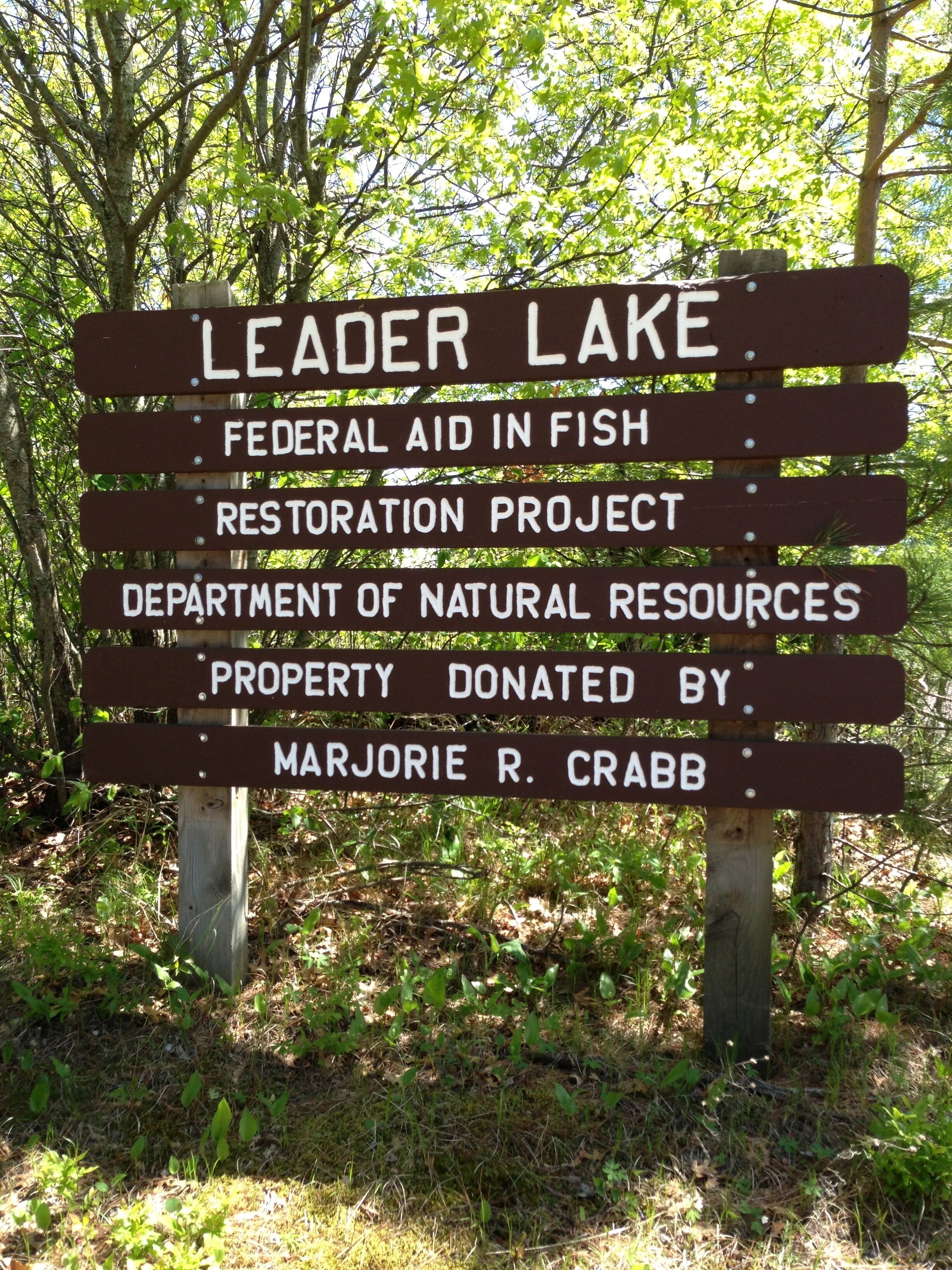 A wooden sign with text about Leader Lake, federal aid in fish restoration, and a donation by Marjorie R. Crabb.