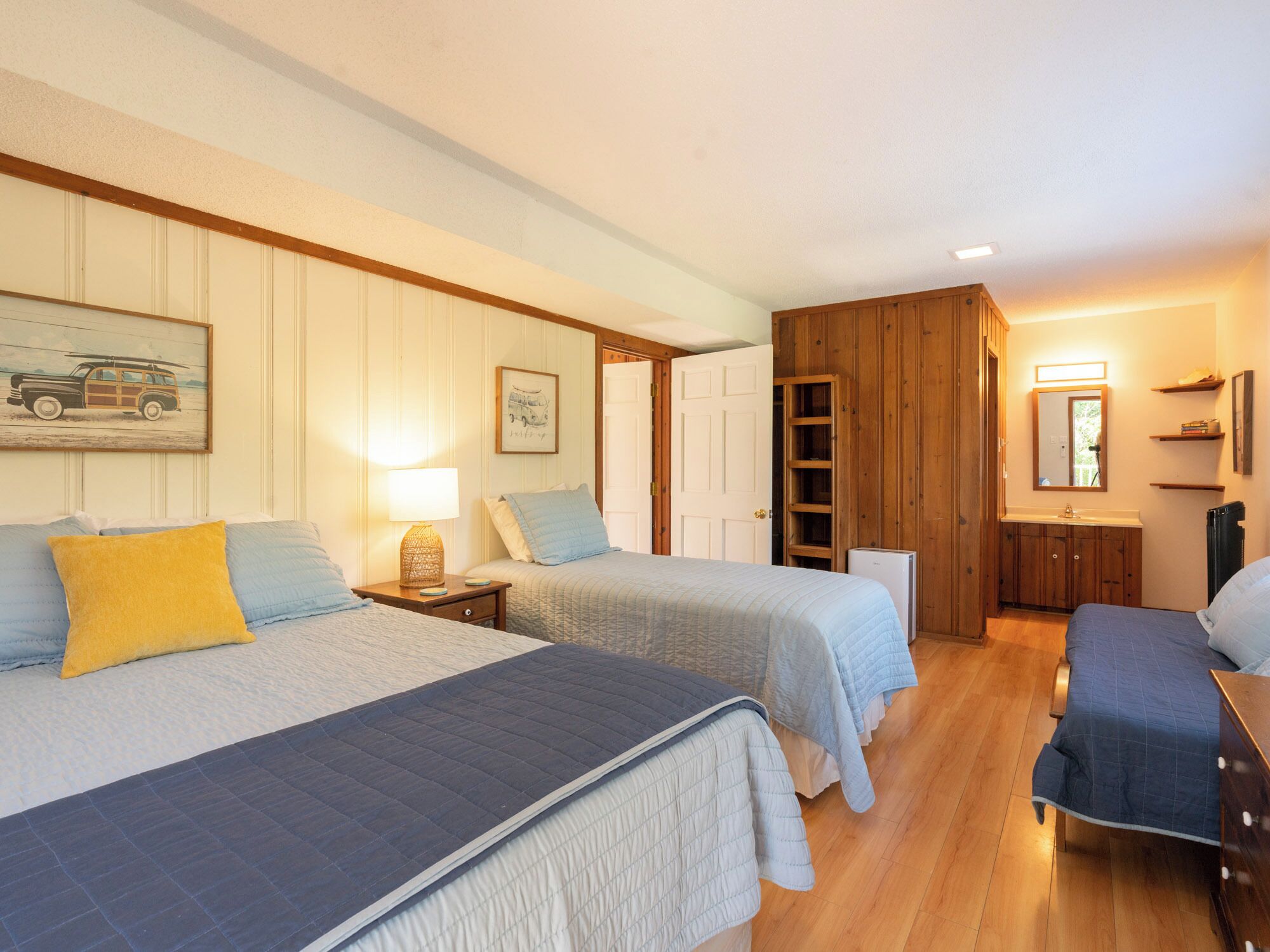 Une chambre avec deux lits, une table de chevet avec une lampe, une commode, un miroir et une porte donnant sur une autre pièce.