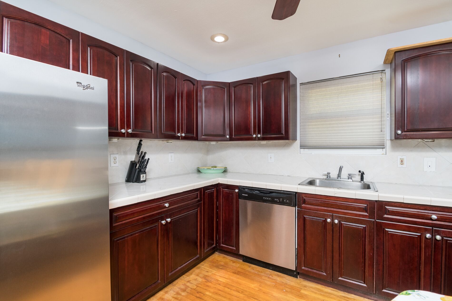 A kitchen with dark wooden cabinets, a stainless steel refrigerator, a dishwasher, and a window with blinds.
