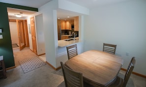 Een eetgedeelte met een houten tafel en stoelen, een keuken met houten keukenkastjes en een deur naar een andere kamer.