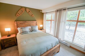 A bedroom with a wooden headboard, a large bed with multiple pillows, bedside tables with lamps, and a window with blinds. - Stoney Creek Northstar 14 (Whistler)