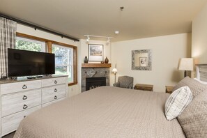 A bedroom with a large bed, a flat-screen TV on a dresser, a fireplace, and a window with curtains.