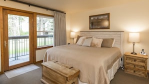 A bedroom with a large bed, two bedside tables with lamps, a wooden bench, and a sliding glass door leading to an outdoor area.