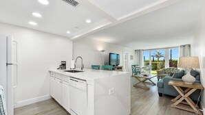 Uma cozinha moderna com uma ilha central, uma sala de jantar com televisão e uma sala de estar com sofá e abajur.