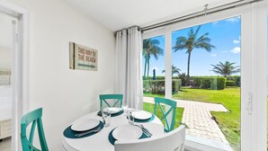 Uma área de refeições com uma mesa posta para dois, um letreiro na parede e vista para um gramado e palmeiras através de grandes janelas.