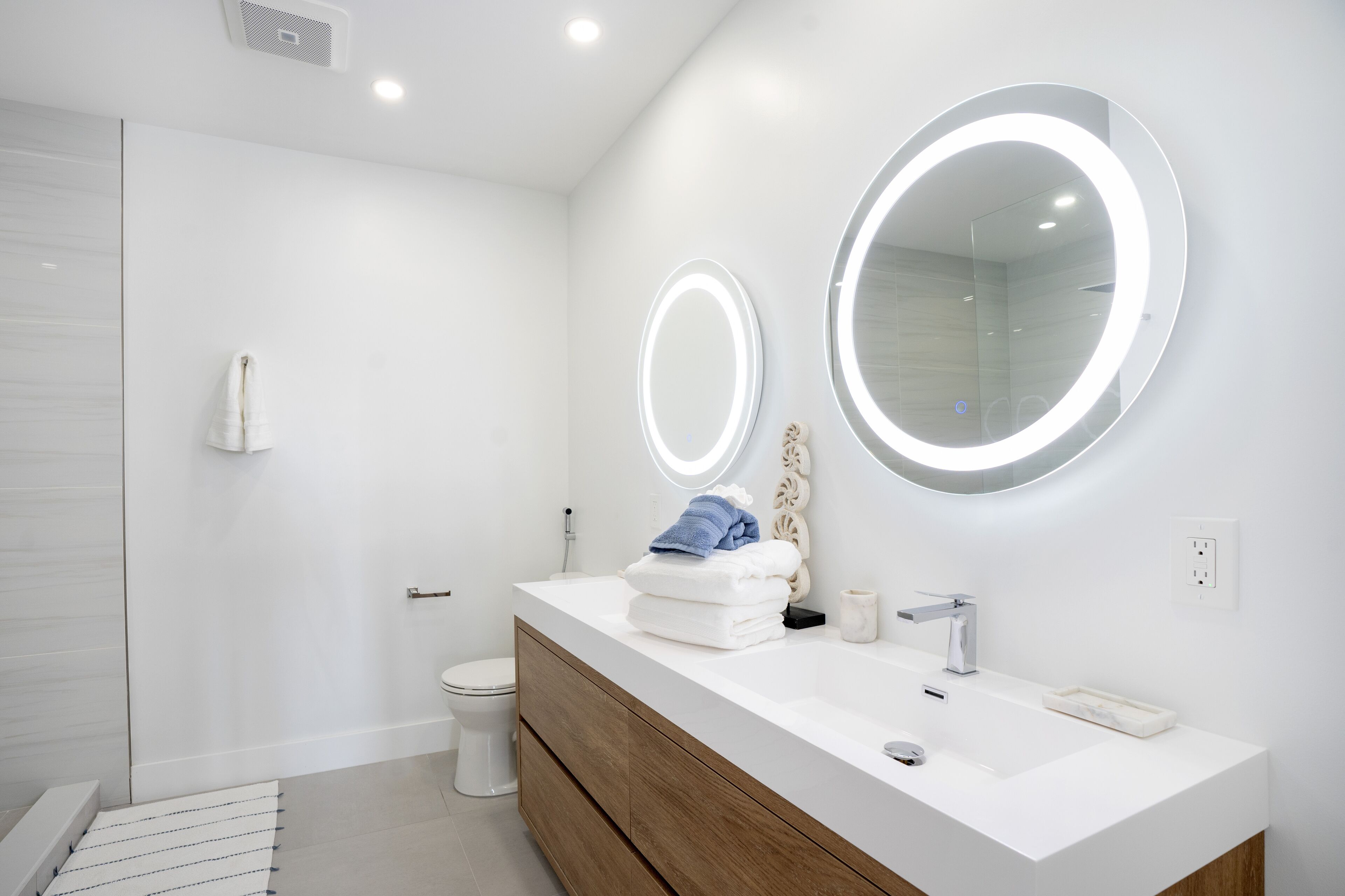 A modern bathroom with a white vanity, two illuminated mirrors, and a shower area.