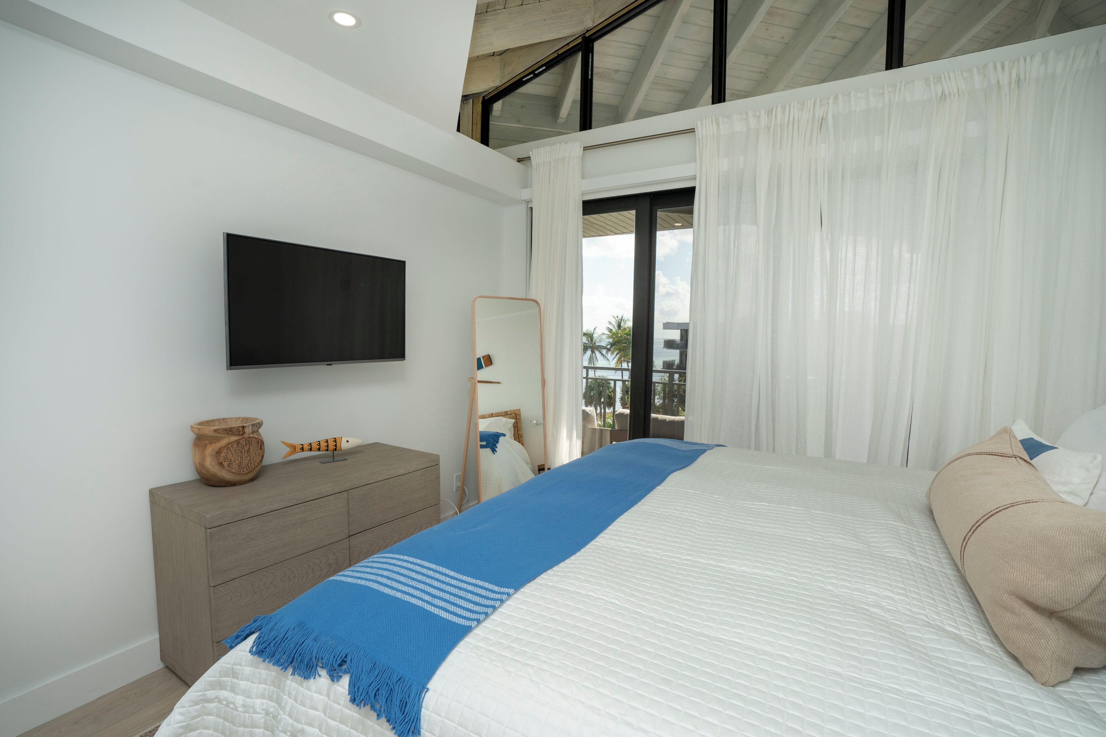 A modern bedroom with a large bed, a flat-screen TV, a wooden dresser, and a view of the outdoors through large windows.