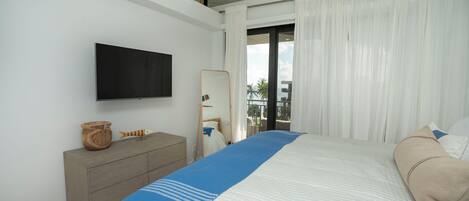 A modern bedroom with a large bed, a flat-screen TV, a wooden dresser, and a view of the outdoors through large windows.