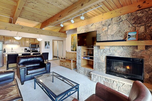 A living room with a stone fireplace, leather sofas, a glass coffee table, wooden beams, and a kitchen in the background.