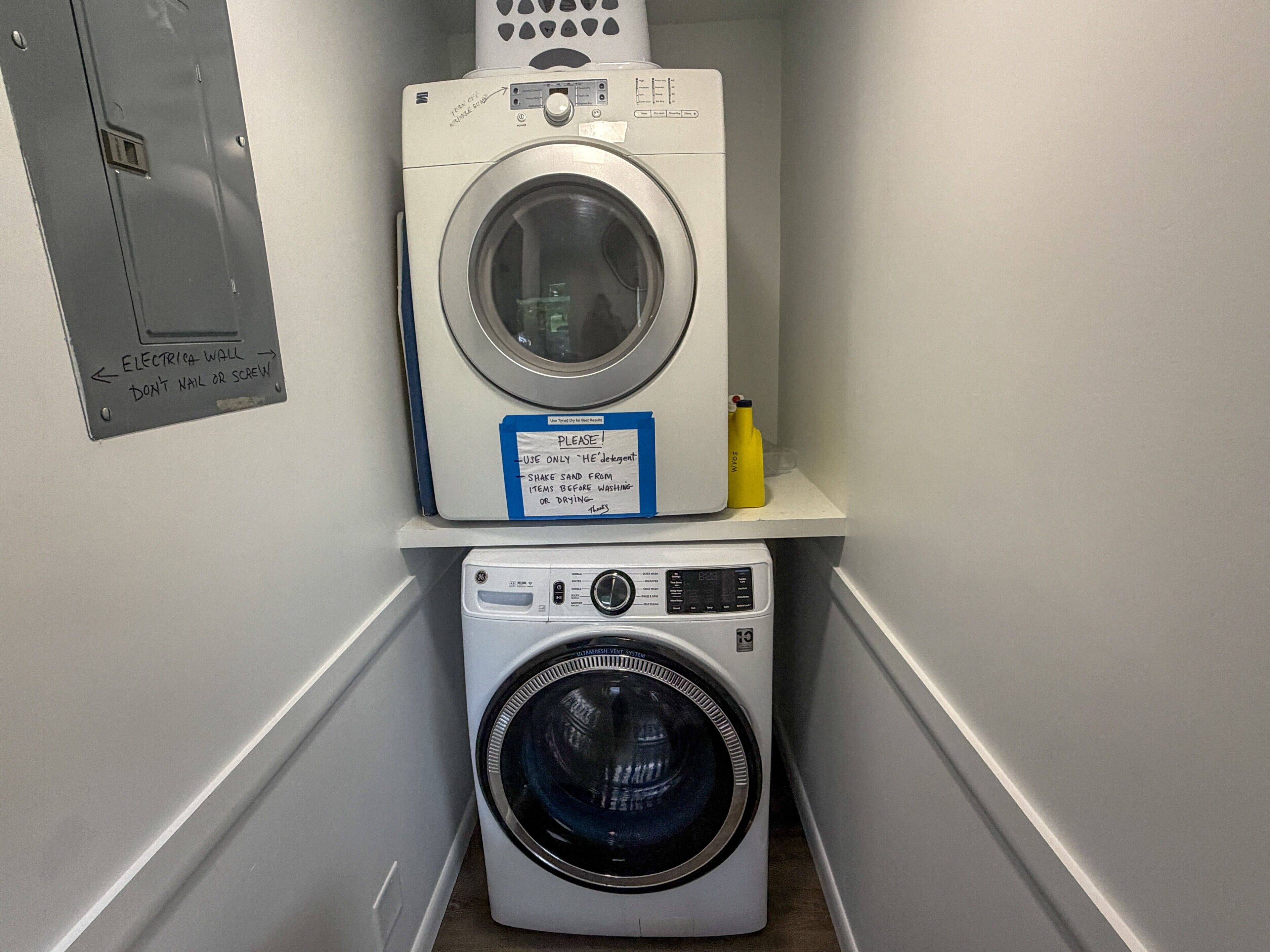 A laundry area with two stacked washing machines, a sign instructing to use only 'he detergents', and a yellow bottle on the shelf.