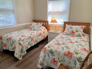 A bedroom with two single beds, each with matching floral bedding, a nightstand with a lamp, and a window with blinds.
