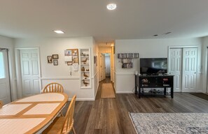 A dining area with a wooden table and chairs, a hallway with a rug, and a living room with a TV and bookshelves.