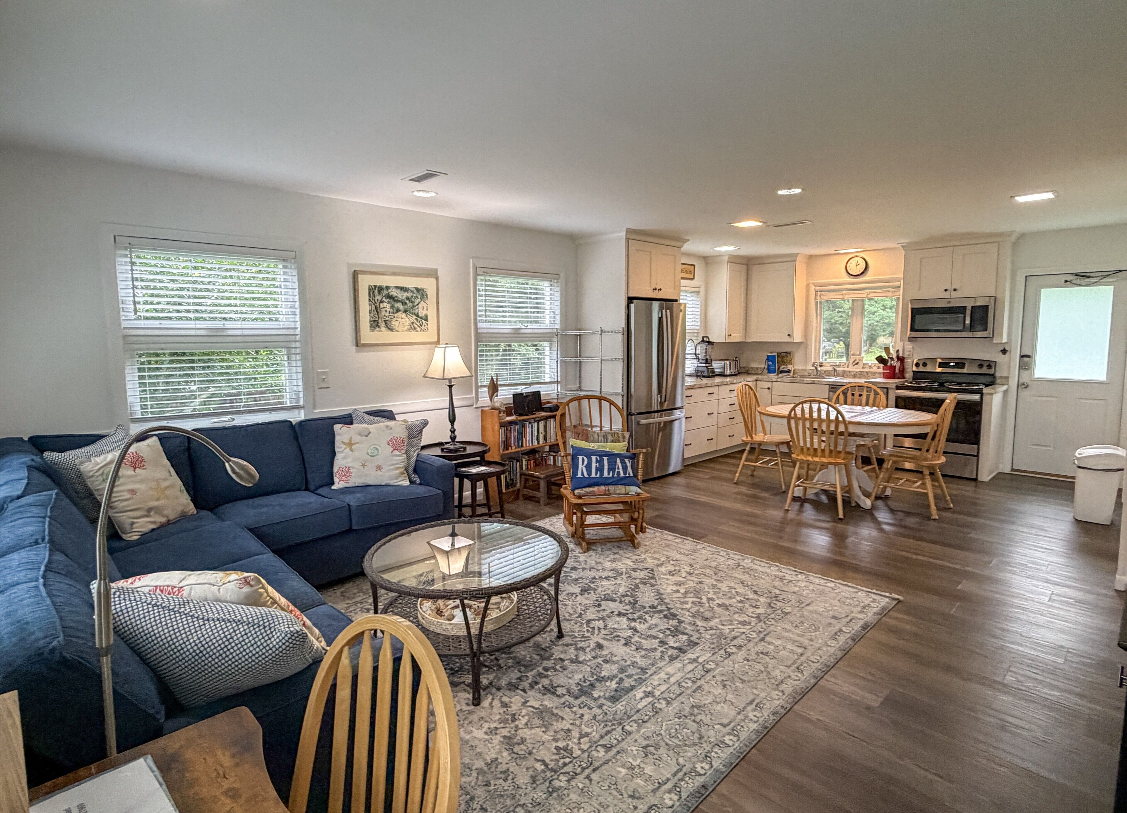 A living room with a blue sofa, a coffee table, and a rug. There is a kitchen area with a refrigerator, microwave, and stove.