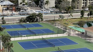 Un complexe de courts de tennis comprenant plusieurs terrains, entouré d’une clôture et situé à proximité d’une route où sont garées des voitures et des bateaux.