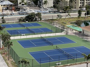 A tennis court complex with multiple courts, surrounded by a fence, and adjacent to a road with parked cars and boats. - Gorgeous Beach Front! Pool, Tennis, Pickleball, Wi-Fi, Extra special Unit!! (Fort Myers Beach)