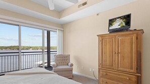 A bedroom with a large bed, a wooden cabinet, a television, a chair, and a balcony with a view of the water.