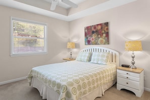 A bedroom with a bed, two bedside tables, a ceiling fan, a window with blinds, and a floral painting on the wall.