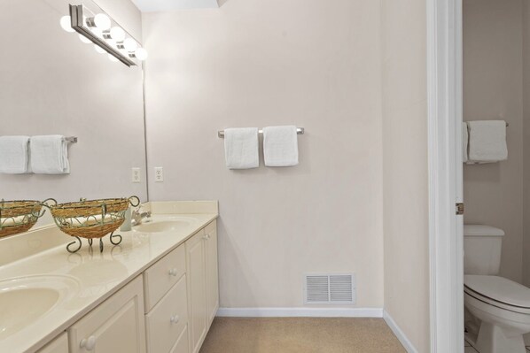 A bathroom with a double sink, two wicker baskets, a mirror with lights, and a toilet.