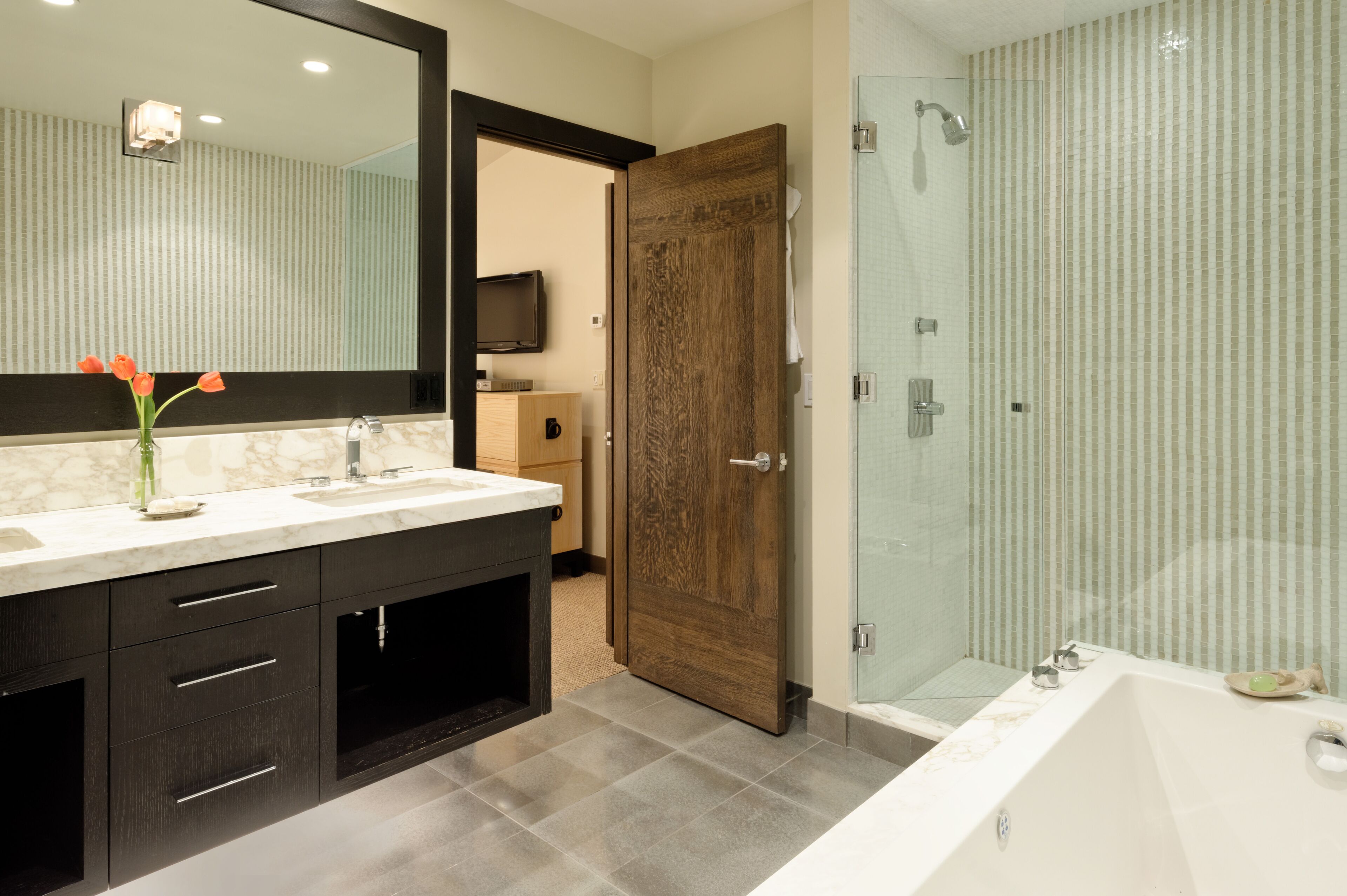 Une salle de bain moderne dotée d’un grand miroir, de meubles de rangement aux couleurs sombres, d’un comptoir en marbre et d’une cabine de douche en verre.