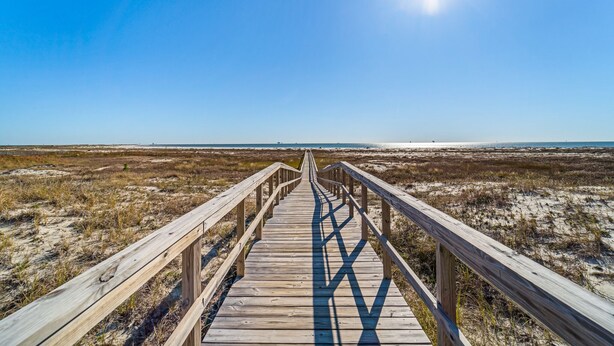 Ein hölzerner Steg führt zu einem Strand bei blauem Himmel und Sonnenschein.