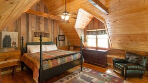 Un dormitorio acogedor en el ático, con techo inclinado de madera, una cama con colcha colorida, un escritorio con lámpara y un sillón de cuero.