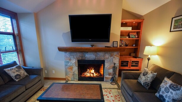 A living room with a stone fireplace, a flat-screen TV mounted above it, a sofa, a coffee table, and a bookshelf.