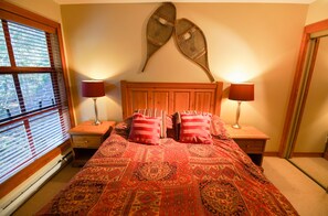 A bedroom with a wooden headboard, two bedside tables with lamps, a patterned bedspread, and a window with blinds.