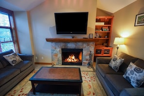Ein Wohnzimmer mit Kamin aus Stein, einem darüber montierten Flachbildfernseher, einer Couch, einem Couchtisch und einem Bücherregal.