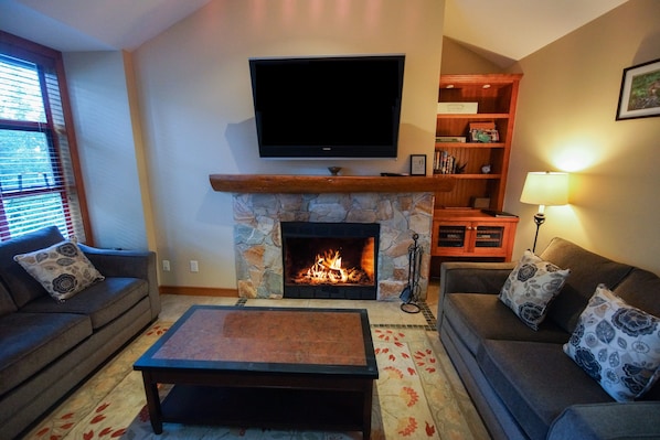 A living room with a stone fireplace, a flat-screen TV mounted above it, a sofa, a coffee table, and a bookshelf. - The Woods - Renovated, Family Friendly Condo on Free Shuttle and Golf Course (Whistler)