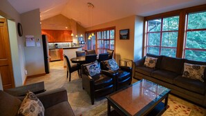 A spacious living area with a sofa, armchairs, and a coffee table. A kitchen is visible in the background with wooden cabinets and a dining area with chairs and a table.