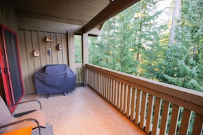 A covered outdoor area with a barbecue grill, a chair, and a wooden railing overlooking a forest. - The Woods - Renovated, Family Friendly Condo on Free Shuttle and Golf Course (Whistler)