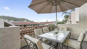A terrace with a table and chairs, an umbrella, and a view of buildings and hills. - C54: 2 Bedroom, 2 Bath Condo, Balcony with Views, Pool, BBQ (Avalon)