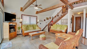 Ein geräumiges Wohnzimmer mit hohen Decken, freiliegenden Holzbalken, einem Fernseher und zwei Sofas mit gemusterten Kissen.