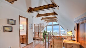Ein geräumiges Dachzimmer mit Holzbalken, einem großen Fenster mit Meerblick und einer gemütlichen Sitzecke mit Schreibtisch und Lampe.